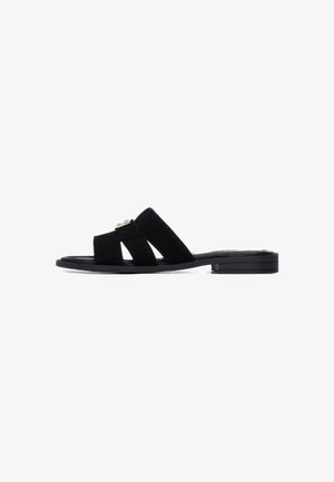 Sandalias tipo "slide" de ante negro con suela plana. Presentan dos tiras anchas que se superponen y un adorno decorativo en la parte delantera. Diseño simple y minimalista.