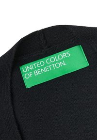 Černá pletená látka se zelenou štítkem s bílým nápisem „UNITED COLORS OF BENETTON“. Textura působí měkce a jemně pleteně.