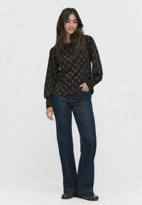 Schwarzes langärmliges Oberteil mit sich überlappendem kreisförmigen Muster in Orange, kombiniert mit weit geschnittenen dunkelblauen Jeans. Beide Kleidungsstücke haben eine glatte Textur.