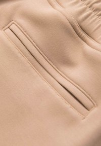 Tissu beige en gros plan montrant une poche fendue sur le côté avec une couture soignée et une partie d'une ceinture élastique sur un vêtement.