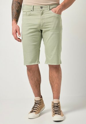 Mann trägt hellgrüne knielange Shorts und beige High-Top-Sneakers mit schwarzen Schnürsenkeln, steht mit einer Hand in der Tasche.