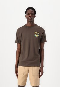 Dunkelbraunes Baumwoll-T-Shirt mit geripptem Rundhalsausschnitt, auf der linken Brust mit einem bunten Blumenmotiv. Getragen mit beigen Shorts.