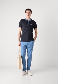 Mornarsko modra polo majica z svetlo modrim ovratnikom in manšeti, v kombinaciji s svetlo modrimi hlačami in belimi športnimi copati. Model drži bež torbo.