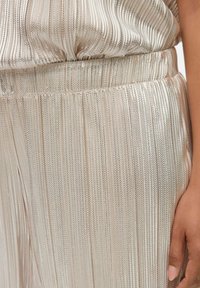 Tissu plissé doré métallisé avec une texture côtelée, doté d'une taille élastique. La lumière se reflète sur la surface lisse, créant des reflets brillants.