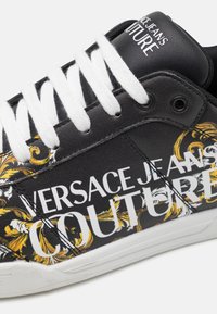 Czarne sneakersy z białymi sznurowadłami i białą gumową podeszwą, zdobione złotymi kwiatowymi wzorami oraz dużym napisem "VERSACE JEANS COUTURE".