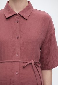 Κοντινή λήψη ενός κοντομάνικου φορέματος πουκάμισο σε μωβ απόχρωση με υφή, γιακά, κουμπιά μπροστά και ζώνη στη μέση που δένει.