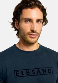 Marineblaues Baumwoll-T-Shirt mit kurzen Ärmeln, das ein markantes schwarzes "ELBSAND"-Logo auf der Brust zeigt. Rundhalsausschnitt, weiche Textur.