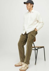 Light striped button-up shirt, olive corduroy cargo pants, and beige sneakers with cream accents. Hat has a black logo. Seated on a chair.