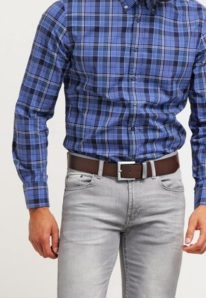 Man met een blauw-zwart geruit overhemd dat is ingestopt in lichtgrijze spijkerbroek met een bruine leren riem.