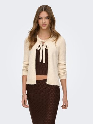 Jeune femme portant une tenue texturée marron en deux parties et un cardigan beige noué par un nœud au niveau du col, debout devant un fond clair.
