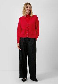 Rote Strickjacke mit V-Ausschnitt, Knopfverschluss und zwei aufgesetzten Taschen, kombiniert mit schwarzen, weit geschnittenen, glitzernden Hosen und schwarzen Schuhen.