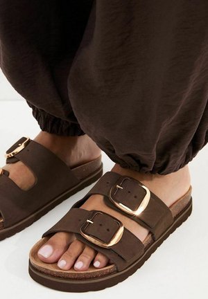 Piedi che indossano sandali marroni con grandi fibbie dorate e pantaloni marroni larghi su una superficie bianca.