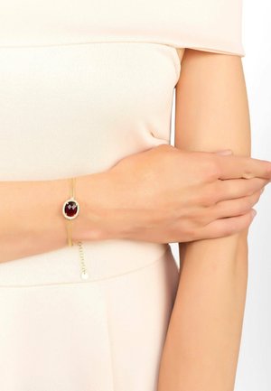 Gold bracelet featuring a large oval red stone surrounded by clear stones, with a two-strand design and an adjustable chain fastener.