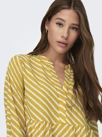 Žuta haljina s bijelim cik-cak prugama. Ima V-izrez i duge rukave. Mekana tkanina, lepršavi dizajn i detalj s nabranim strukom.