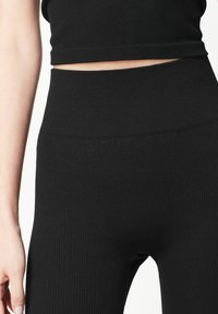 Gros plan sur une personne portant un legging noir à taille haute et côtelé, ainsi qu'un crop top assorti, montrant le torse et le bras gauche.