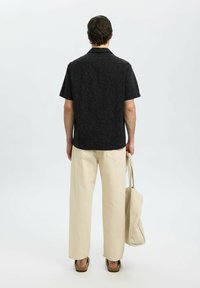 Chemise noire à manches courtes avec un motif de vagues texturé, associée à un pantalon large beige et portant un sac fourre-tout beige. Des chaussures décontractées sont également incluses.