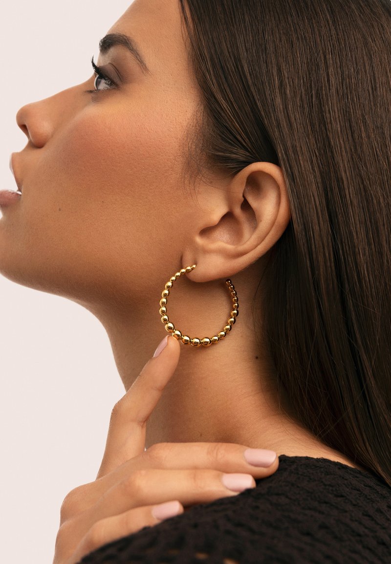 Boucles d'oreilles en or en forme de créole avec un design texturé comportant de petites perles sphériques uniformément espacées, portées à l'oreille, mettant en valeur leur forme circulaire.