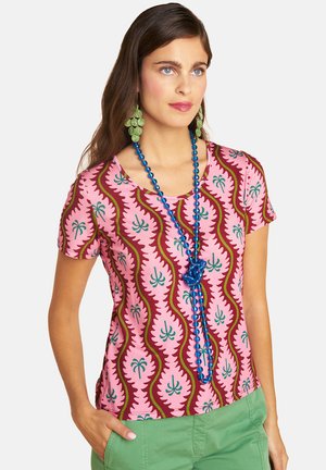 Woman with long brown hair wears a pink patterned shirt, green pants, green leaf earrings, and a long blue beaded necklace.