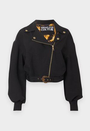 Veste noire courte avec fermeture éclair dorée, boutons et boucle de ceinture, manches longues bouffantes, et étiquette "Versace Jeans Couture" à l'intérieur du col.
