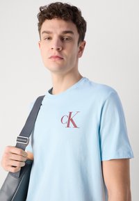 Světle modré bavlněné tričko s vínovým logem "cK" na levém prsu. Model drží šedou tašku přes rameno s popruhem.