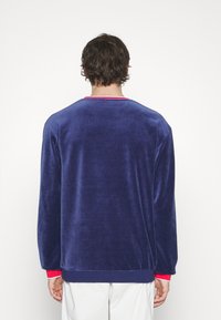 Pulover z navy modrého sametu s dlouhými rukávy, kulatým výstřihem a červenými akcenty na límci a manžetách. Měkká textura a volný střih.