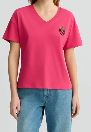 Camiseta de algodón rosa con escote en V, mangas cortas y una aplicación de fresa en el lado izquierdo del pecho. Corte informal, textura suave y diseño simple.