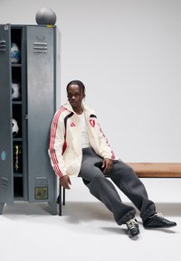 White jacket with red accents, gray sweatpants, and black sneakers. Background features a gray locker and sports equipment.