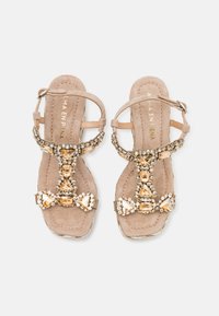 Sandales en suède beige avec un design à T, ornées de décorations en or et en pierres gemmes transparentes, une bride ajustable à la cheville et une semelle intérieure rembourrée.