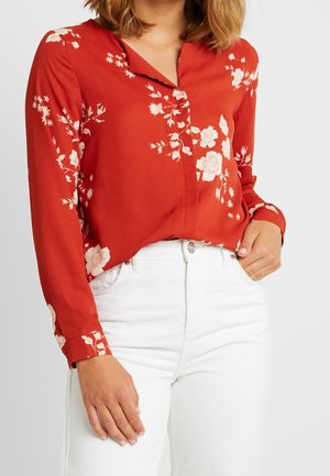 Kvinde iført en rød bluse med lange ærmer med hvide blomsterprint, stoppet ned i hvide bukser med høj talje, ene hånd i lommen.
