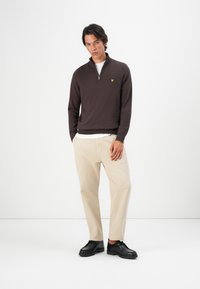 Brun zip-genser, beige bukser og svarte sko. Genseren har en gul logo på brystet og en teksturert strikk.