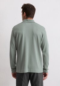 Polo cu mânecă lungă, de culoare verde deschis, realizat dintr-un material moale. Dispune de un guler clasic și manșete ribbate, cu o croială lejeră și fără logo-uri vizibile.