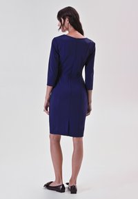 Navyblaues Kleid mit Dreiviertelärmeln, taillierter Passform und Reißverschluss hinten. Verfügt über einen Schlitz hinten für mehr Bewegungsfreiheit. Getragen mit dunklen flachen Schuhen.