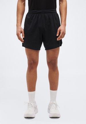 Muž oblečený v obyčajných čiernych športových šortkách, bielych posádkových ponožkách a bielych bežeckých topánkach stojaci pred jednoduchým svetlým pozadím.