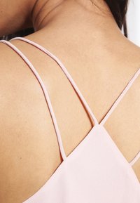 Close-up van de rug van een persoon die een lichtroze top draagt met dunne, dubbelgekruiste schouderbandjes. Huidskleur is lichtbruin.