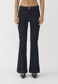 Jeans acampanados de color azul oscuro con una cintura ajustada, bolsillos frontales y costuras en contraste de color naranja. Combinados con tacones negros con tachuelas.