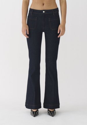 Dark blue flared jeans with a fitted waist, front pockets, and contrasting orange stitching. Paired with black studded heels.