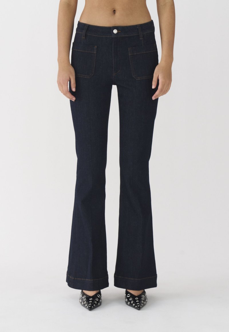 Jeans acampanados de color azul oscuro con una cintura ajustada, bolsillos frontales y costuras en contraste de color naranja. Combinados con tacones negros con tachuelas.
