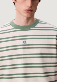 Camisa de manga corta a rayas en color blanco roto con rayas verdes horizontales. Presenta un cuello redondo acanalado verde y un pequeño logo en el pecho.