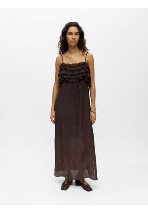 Femme aux longs cheveux bouclés portant une robe maxi marron foncé sans manches avec un haut à volants et des chaussures marron foncé assorties, debout sur fond blanc.
