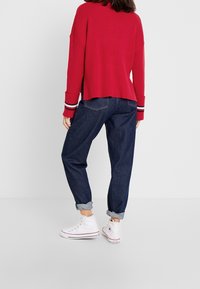 Czerwony sweter z prążkowanego dzianiny z paskowanymi mankietami, zestawiony z luźnymi, ciemnoniebieskimi dżinsami i białymi, wysokimi trampkami, z podwiniętymi rękawami.