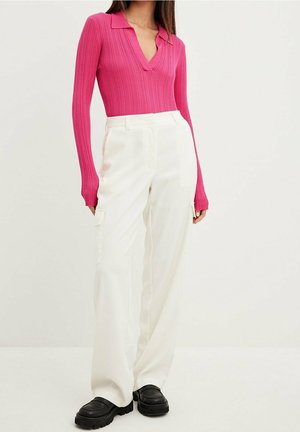 Vrouw draagt een nauwsluitende roze geribbelde top met lange mouwen en kraag, witte broek met hoge taille en brede pijpen en zwarte chunky schoenen, staand tegen een effen achtergrond.