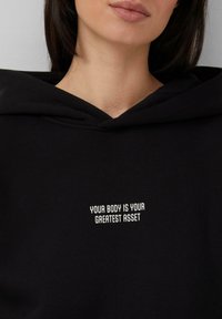 Zwarte hoodie met witte tekst waarop staat "JOUW LICHAAM IS JE GROOTSTE ACTIVUM," met een ruime hals en zachte, gestructureerde stof.