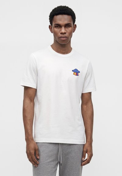 Νέος άνδρας που φοράει λευκό t-shirt με μικρό μπλε UFO στο αριστερό στήθος που εκπέμπει ουράνια τόξα ακτίνες, συνδυασμένο με γκρι φόρμα.