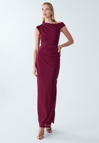 Sieviete stāv un valkā grīdas garuma, bez piedurknēm burgundijas krāsas kleitu ar drapēta kakla izgriezumu un zelta krāsas augstpapēžu sandales.