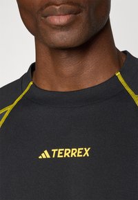 Camisa desportiva preta com detalhes amarelos na gola e no logótipo. O tecido parece suave, com um ajuste justo na área do pescoço e dos ombros.