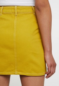 Jupe mini en denim jaune avec un design ajusté, des coutures visibles et des passants de ceinture à la taille. Texture lisse.