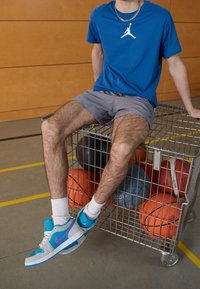 Sneakers med en ljusgrå och blå design, med lila accenter. Modellen bär en blå t-shirt och grå shorts medan han sitter på en vagn med basketballer.