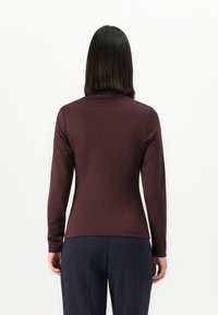 Vrouw met steil donker haar draagt een lange mouwen bordeauxrode top en marineblauwe broek, staande met gezicht naar een effen witte achtergrond.