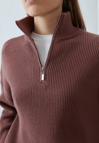 Hnědý žebrovaný pulovr s vysokým límcem a polozipem. Má hladký povrch a stříbrný zip jako akcent.
