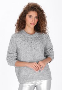 Grå strikket sweater med rund halsudskæring, pyntet med sølvstuder på brystet. Blød tekstur, afslappet pasform og ribstrikkede ærmeafslutninger.